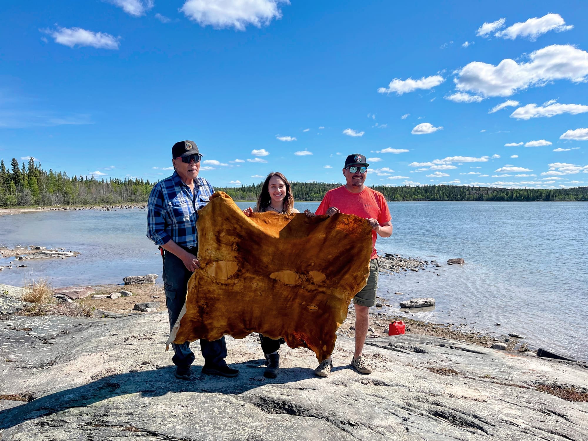 Three Indigenous people hold a tanned hyde in front of a lake.