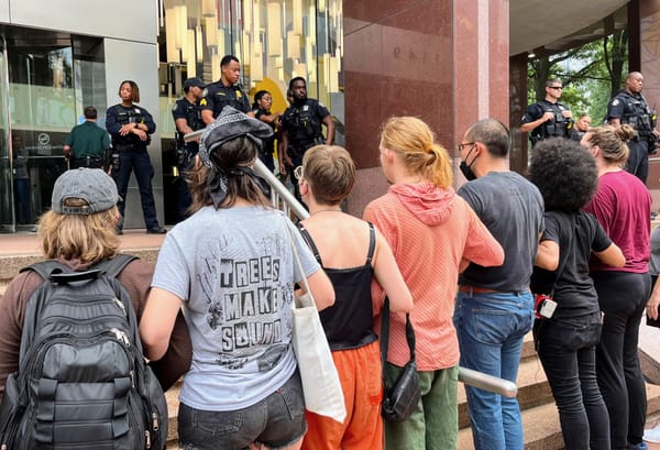 A row of protesters, facing away from the camera, lock arms at the foot of some steps outside of a bank. At the top of the s