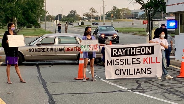 Masked activists hold signs and a banner that reads "Raytheon Missiles Kill Civilians" and use a car to blockade an entrance.