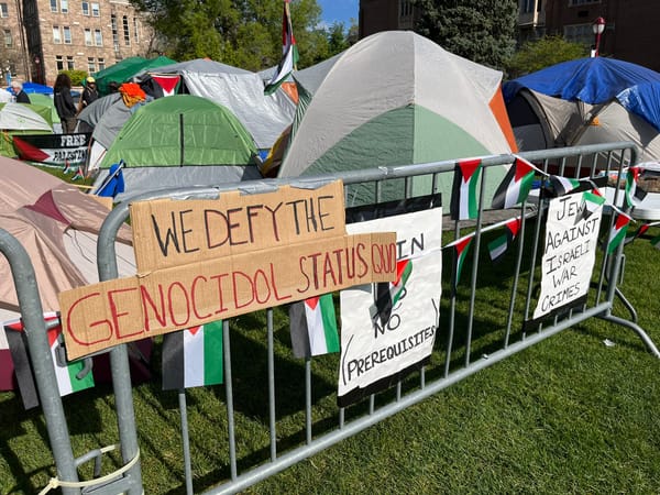 Tents on a campus quad behind a metal barrier lined with Palestinian flags and protest signs. 