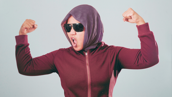 A person wearing sunglasses, a head scarf and a hoodie raises both fists powerfully.